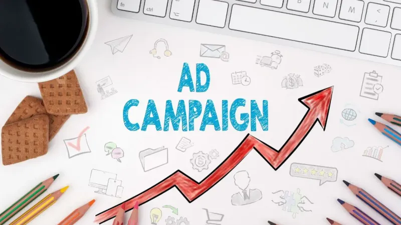 The words Ad Campaign and a rising red arrow drawn on paper, with coloured pencils, a keyboard and a cup of coffee