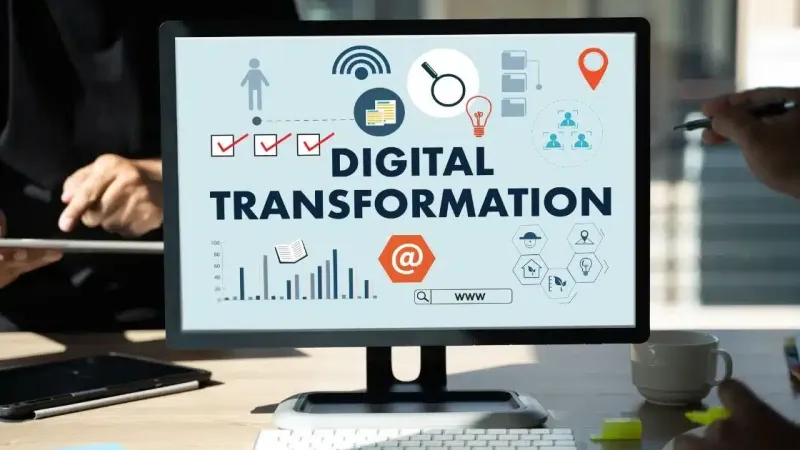 Monitor bertuliskan Digital Transformation dengan ikon digital, di meja kerja dengan tablet dan cangkir kopi