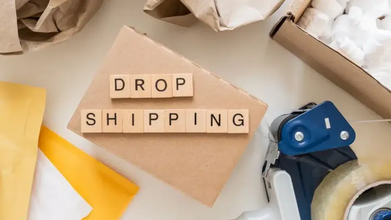 Huruf kayu bertuliskan DROP SHIPPING di atas kardus, dikelilingi amplop, kertas pembungkus, dan dispenser selotip