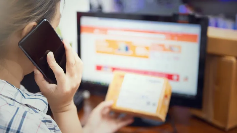 Penjual online menelepon sambil memegang paket di depan monitor yang menampilkan halaman toko online