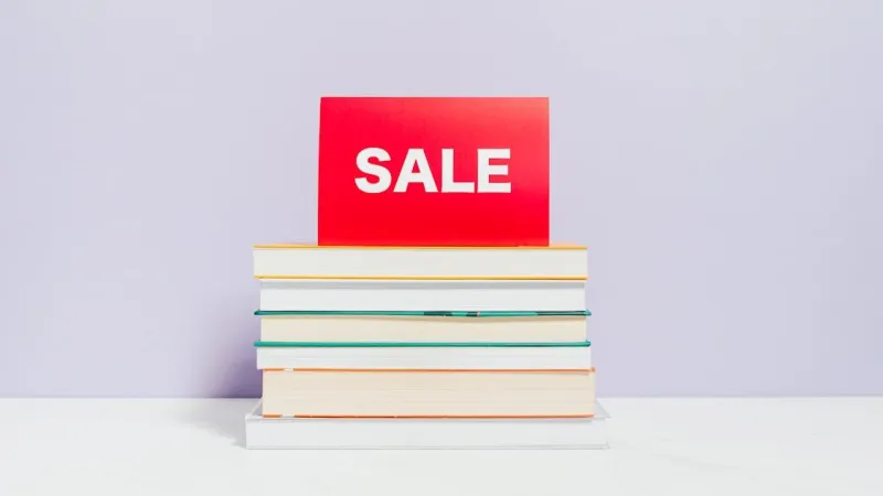 Tumpukan buku dengan kartu merah bertuliskan SALE di atasnya, berlatar dinding ungu muda