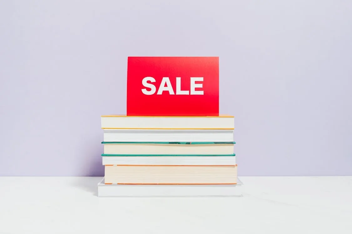 A stack of books topped with a red card reading SALE, against a pale lilac wall