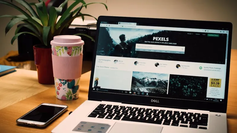 Laptop Dell menampilkan situs Pexels di meja kayu, di samping tumbler bermotif bunga, ponsel, dan tanaman