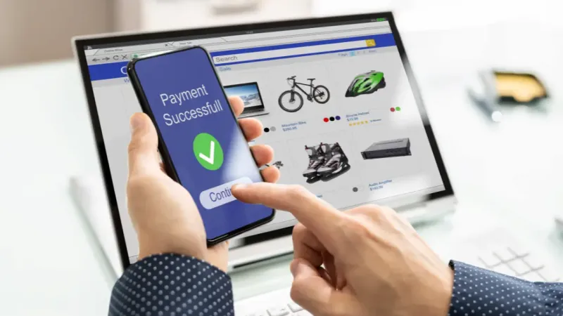 Tangan memegang ponsel bertuliskan pembayaran berhasil di depan laptop yang menampilkan halaman toko online